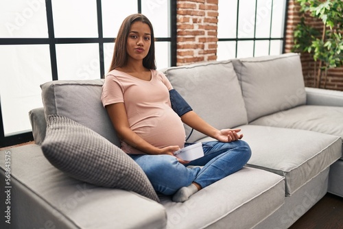 Young pregnant woman using blood pressure monitor sitting on the sofa looking at the camera blowing a kiss being lovely and sexy. love expression.
