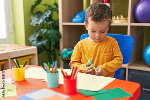 Adorable caucasian boy student cutting paper sitting on table at kindergarten