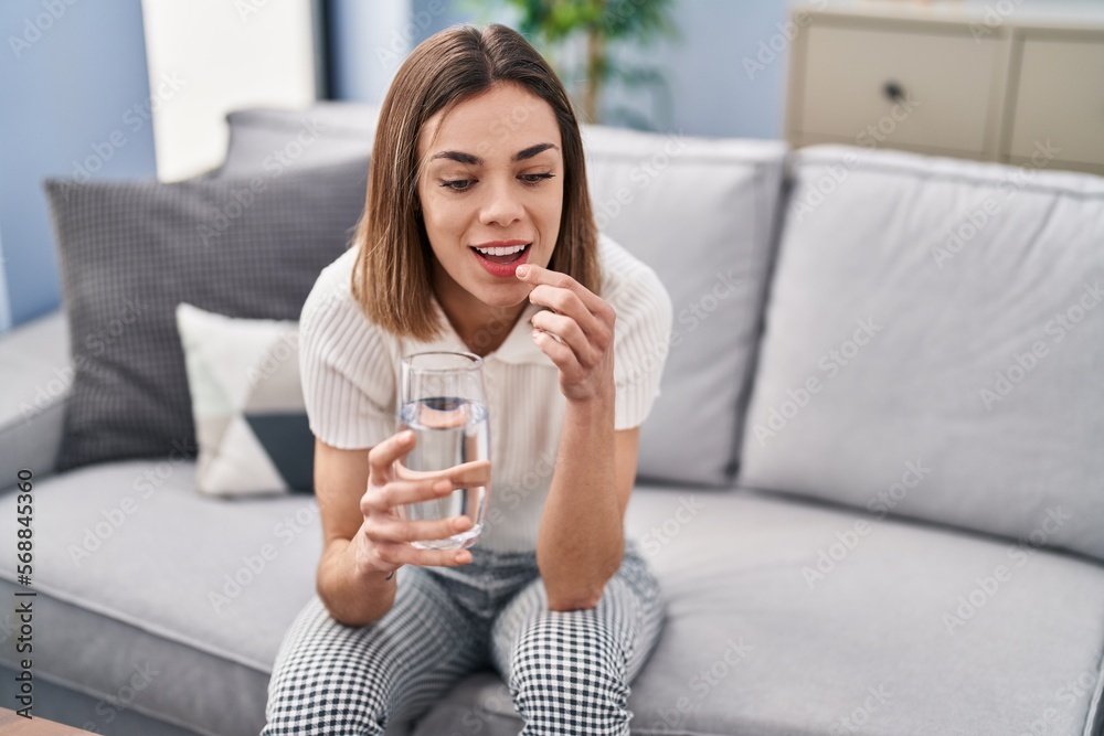 Young beautiful hispanic woman taking pill sitting on sofa at home Young beautiful hispanic woman taking pill sitting on sofa at home