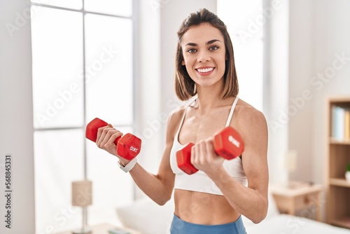 Young beautiful hispanic woman smiling confident using dumbbells training at bedroom