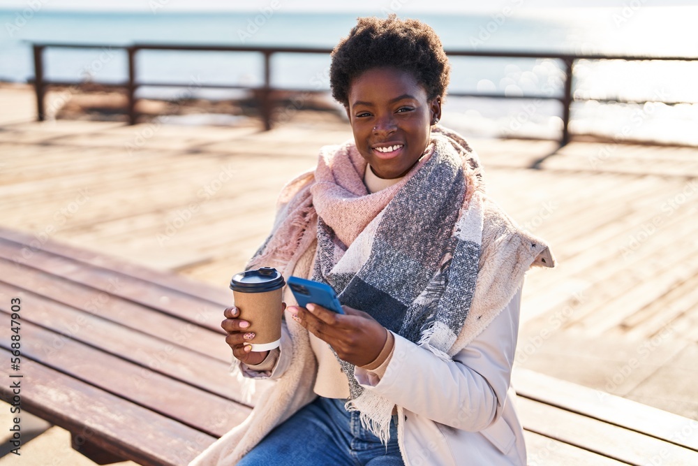Obraz premium African american woman using smartphone drinking coffee sitting on bench at seaside
