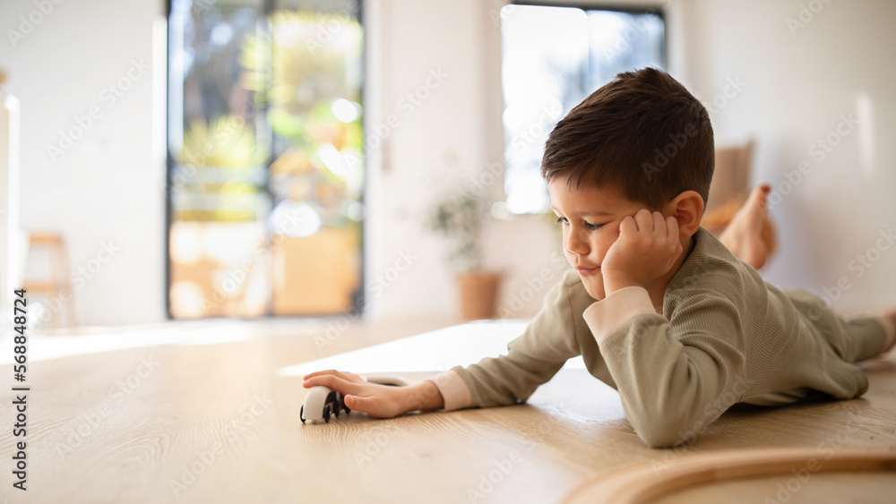 Busy serious bored caucasian little kid lying on floor, playing with toys cars or train, thinking in room interior Busy serious bored caucasian little kid lying on floor, playing with toys cars or train, thinking in room interior