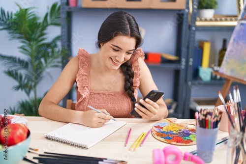 Young hispanic woman artist drawing on notebook using smartphone at art studio