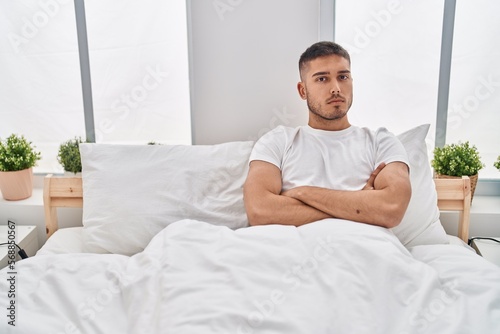 Young hispanic man with serious expression sitting on bed at bedroom