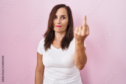 Middle age brunette woman standing over pink background showing middle finger, impolite and rude fuck off expression
