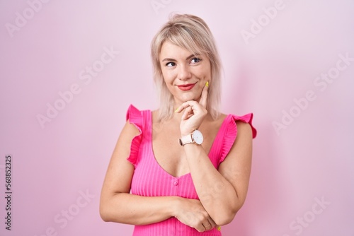 Young caucasian woman standing over pink background with hand on chin thinking about question, pensive expression. smiling and thoughtful face. doubt concept.