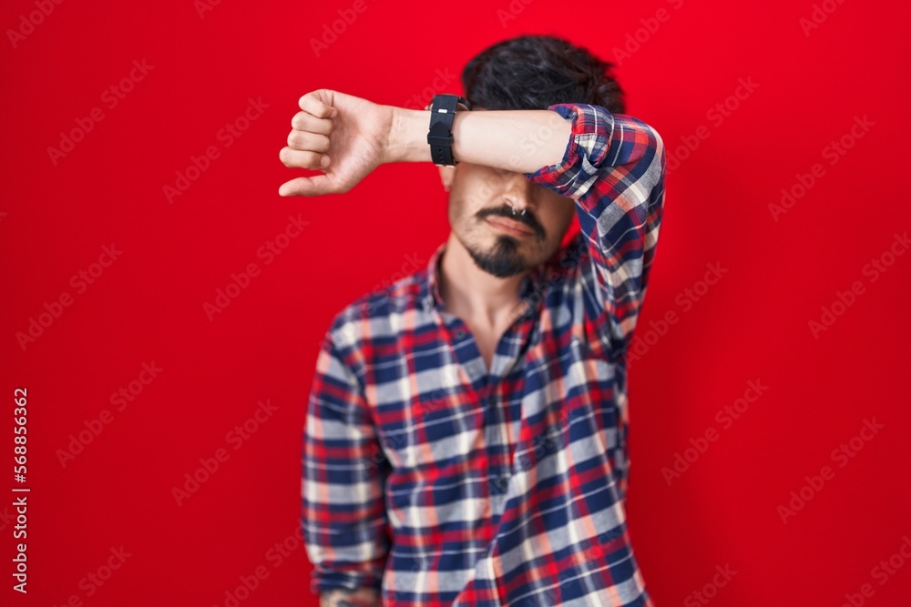 Young hispanic man with beard standing over red background covering eyes with arm, looking serious and sad. sightless, hiding and rejection concept Young hispanic man with beard standing over red background covering eyes with arm, looking serious and sad. sightless, hiding and rejection concept