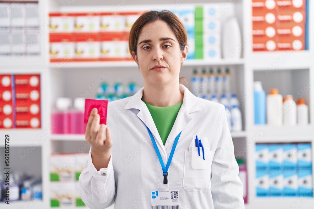 Brunette woman working at pharmacy drugstore holding condom thinking attitude and sober expression looking self confident