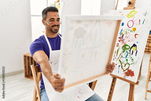 Young hispanic man smiling confident holding draw canvas at art studio