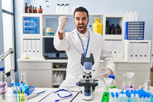 Young hispanic man with beard working at scientist laboratory angry and mad raising fist frustrated and furious while shouting with anger. rage and aggressive concept.