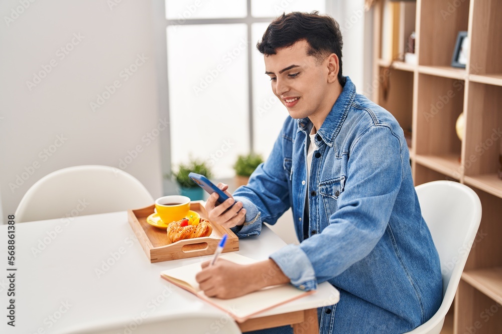 Young non binary man using smartphone and writing on notebook having breakfast at home Young non binary man using smartphone and writing on notebook having breakfast at home