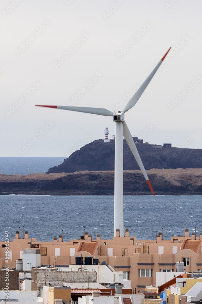 Wind turbine located in small town near blue rippling sea against lighthouse on hill