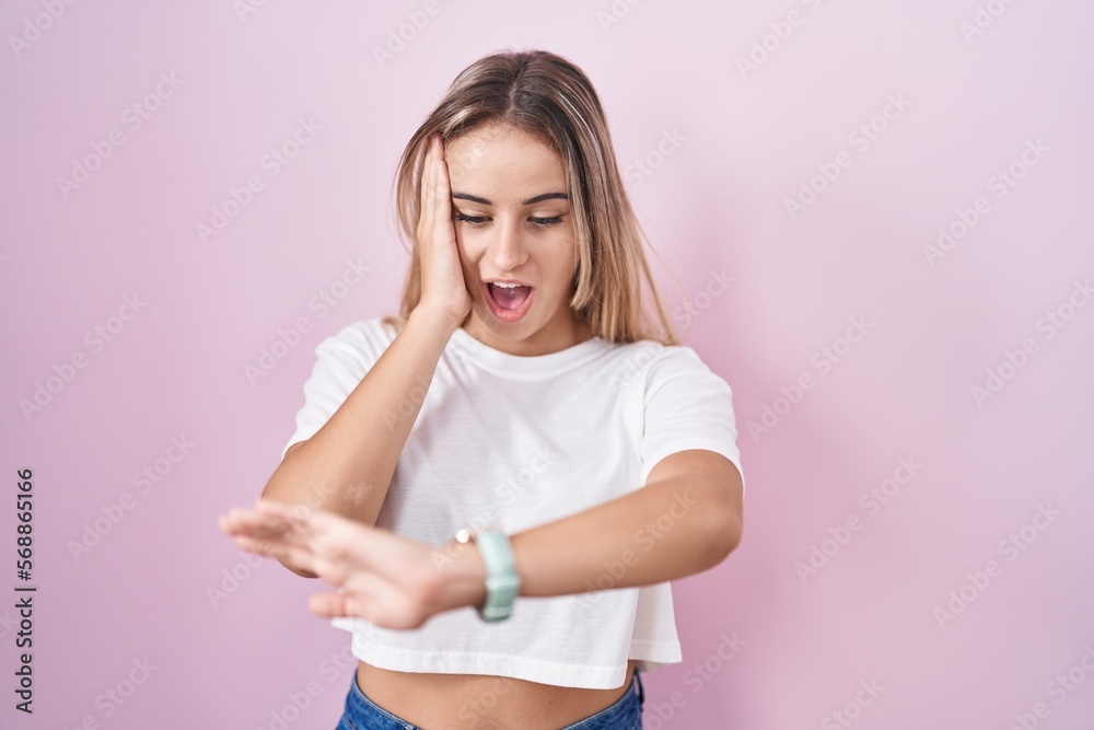 Young blonde woman standing over pink background looking at the watch time worried, afraid of getting late
