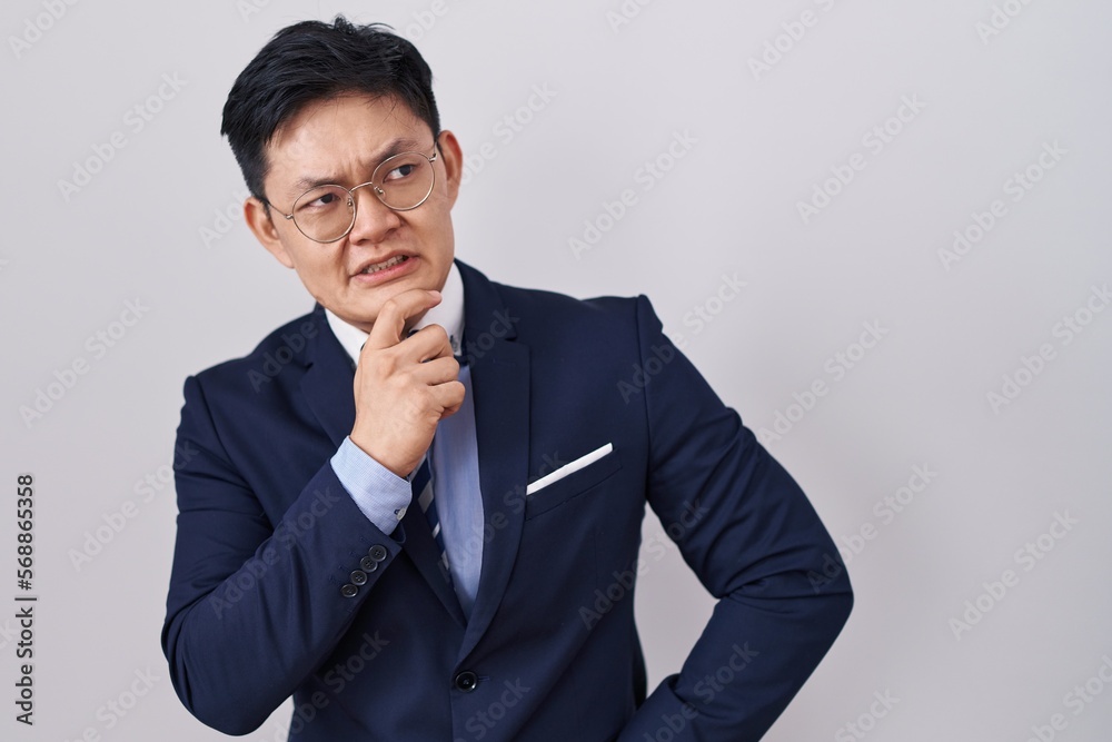 Young asian man wearing business suit and tie thinking worried about a question, concerned and nervous with hand on chin Young asian man wearing business suit and tie thinking worried about a question, concerned and nervous with hand on chin