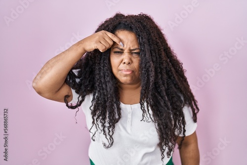 Plus size hispanic woman standing over pink background pointing unhappy to pimple on forehead, ugly infection of blackhead. acne and skin problem