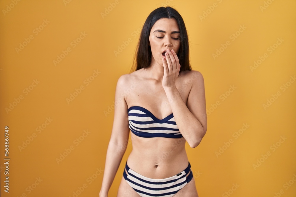 Fototapeta premium Young brunette woman wearing bikini over yellow background bored yawning tired covering mouth with hand. restless and sleepiness.