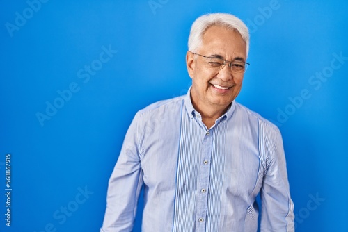 Hispanic senior man wearing glasses winking looking at the camera with sexy expression, cheerful and happy face.