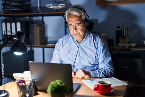 Hispanic senior man wearing call center agent headset at night skeptic and nervous, frowning upset because of problem. negative person.