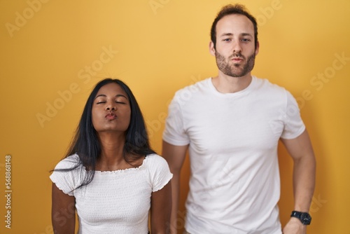 Interracial couple standing over yellow background looking at the camera blowing a kiss on air being lovely and sexy. love expression.