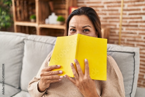 Middle age hispanic woman covering face with book sitting on sofa at home