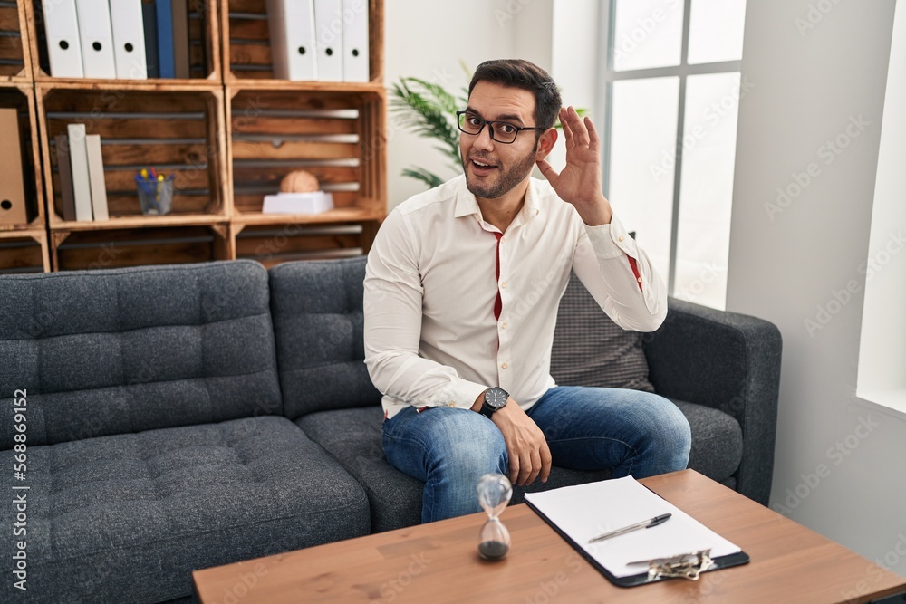 Young hispanic man with beard working at consultation office smiling with hand over ear listening an hearing to rumor or gossip. deafness concept. Young hispanic man with beard working at consultation office smiling with hand over ear listening an hearing to rumor or gossip. deafness concept.