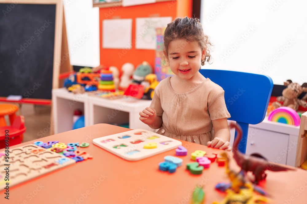Fototapeta premium Adorable hispanic girl playing with maths puzzle game sitting on table at kindergarten