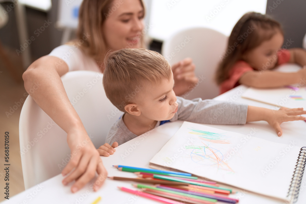 Teacher with boy and girl sitting on table drawing on paper at kindergarten Teacher with boy and girl sitting on table drawing on paper at kindergarten