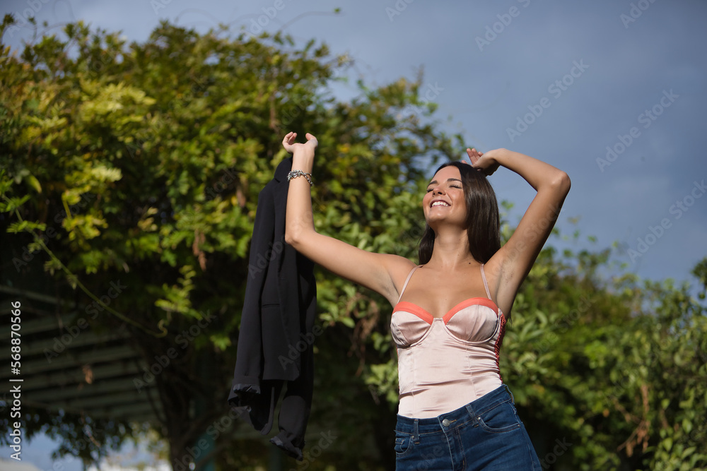 Young and beautiful woman with straight brown hair, orange top, jeans and jacket in hand, arms up smiling and happy. Concept fashion, beauty, trend, relax, millennial.