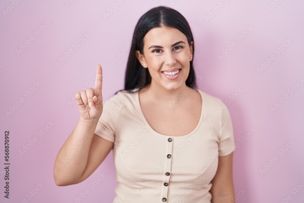 Young hispanic woman standing over pink background showing and pointing up with finger number one while smiling confident and happy. Young hispanic woman standing over pink background showing and pointing up with finger number one while smiling confident and happy.