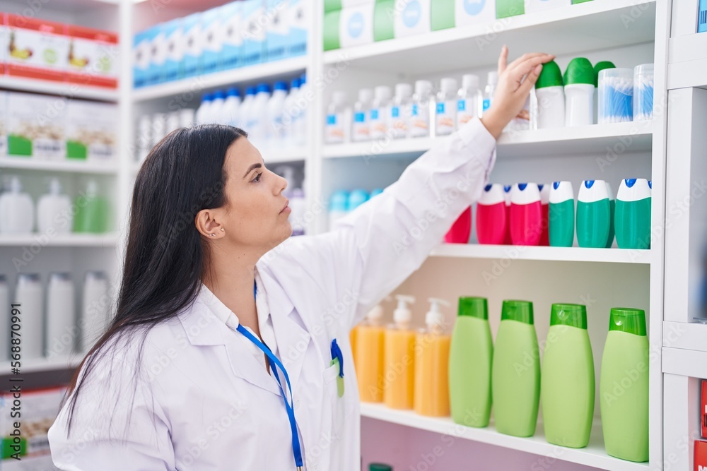 Young beautiful hispanic woman pharmacist holding product on shelving at pharmacy