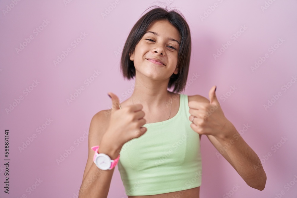 Young girl standing over pink background success sign doing positive gesture with hand, thumbs up smiling and happy. cheerful expression and winner gesture. Young girl standing over pink background success sign doing positive gesture with hand, thumbs up smiling and happy. cheerful expression and winner gesture.