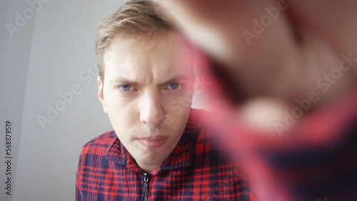 A man in a red shirt is sitting in front of the camera. The young man noticed dirt on the webcam lens. He wipes the camera lens with the cuff of his shirt. View from the laptop webcam.
