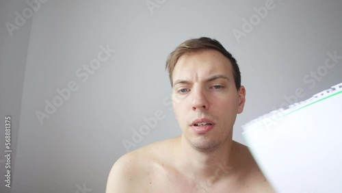 A young man looks into a laptop camera, listens to a video conference. The man sweats, suffers from the heat, fanning himself with a notebook like a fan. View from the laptop webcam.