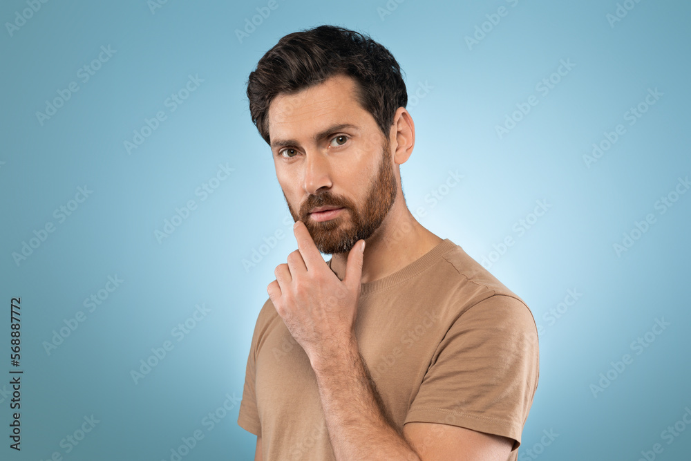 Portrait of middle aged bearded man posing and touching chin, standing over blue studio background Portrait of middle aged bearded man posing and touching chin, standing over blue studio background