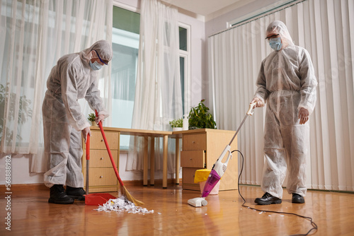 Cleaners in protective suits wiping and vacuum cleaning floor in house of client