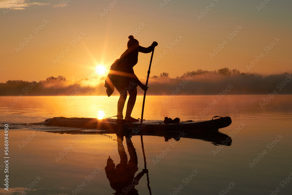 Silhouette of a woman rowing on a SUP during a beautiful autumn sunrise other the misty river