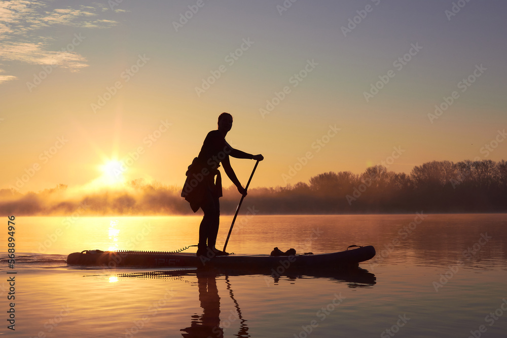 Silhouette of a woman rowing on a SUP during a beautiful autumn sunrise other the misty river