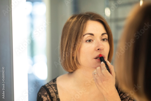 Fashionable beautiful young woman in stylish vintage clothing painting lips with a red lipstick in front of the mirror in lavatory at the night time. Attractive girl preparing for dating and party