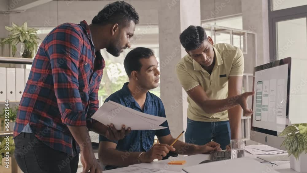 Medium shot of team of Indian UX designers discussing mobile app wireframe on paper and computer screen while working together on project in office of IT company