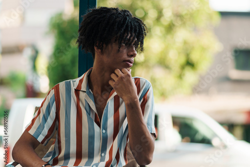 joven afroamericano posando y sentado en la ciudad en un día soleado, mirando hacia un costado con una camisa con líneas de colores, esperando.