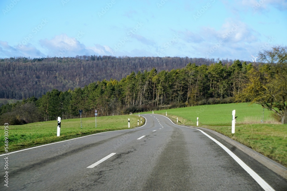 Landschaft Panorama mit kurviger Hauptstraße zwischen grünen Wiesen vor Wald und blauem Himmel mit weißem Wolkengebilde bei Sonne am Nachmittag im Winter