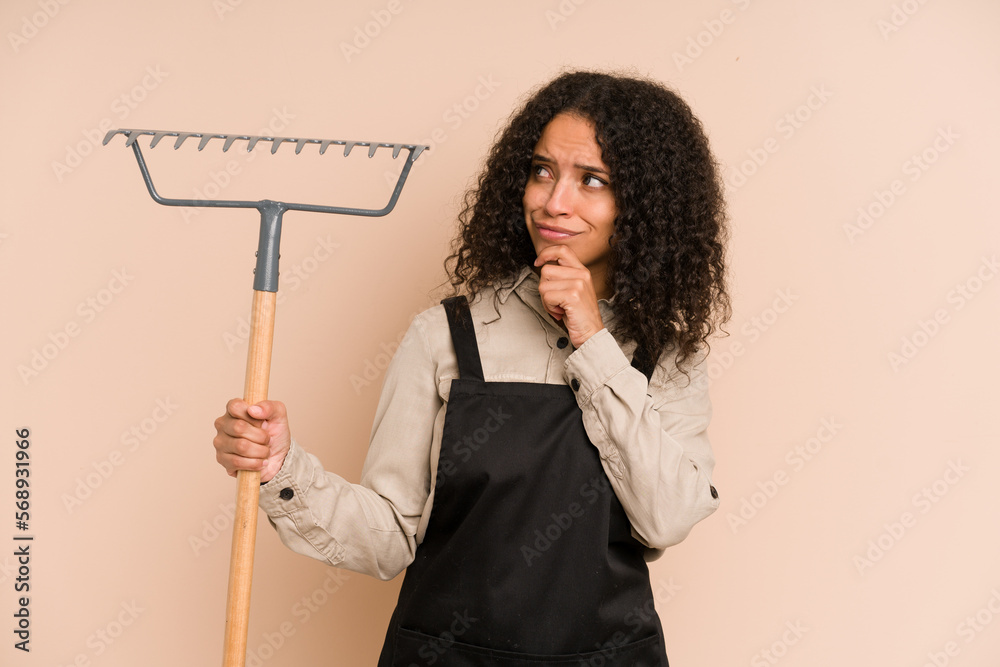 Young african american gardener woman holding a rake isolated looking sideways with doubtful and skeptical expression. Young african american gardener woman holding a rake isolated looking sideways with doubtful and skeptical expression.