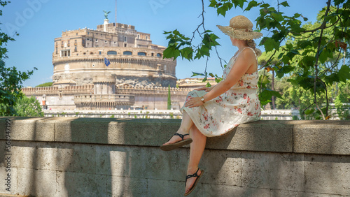 Eine Frau im Sommerkleid sitz auf einer Mauer am Tiber und Blickt zur Engelsburg in Rom, Italien, Europa. photo