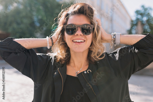 Close up portrait of adorable happy girl with wonderful smile posing to camera while wearing sunglasses and black jacket on background of city square in sunlight 