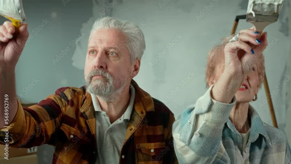 Elderly man and woman are painting glass with white paint using brushes. Gray haired a couple of pensioners ia filling the screen frame with white paint against backdrop of renovation apartment. Elderly man and woman are painting glass with white paint using brushes. Gray haired a couple of pensioners ia filling the screen frame with white paint against backdrop of renovation apartment.
