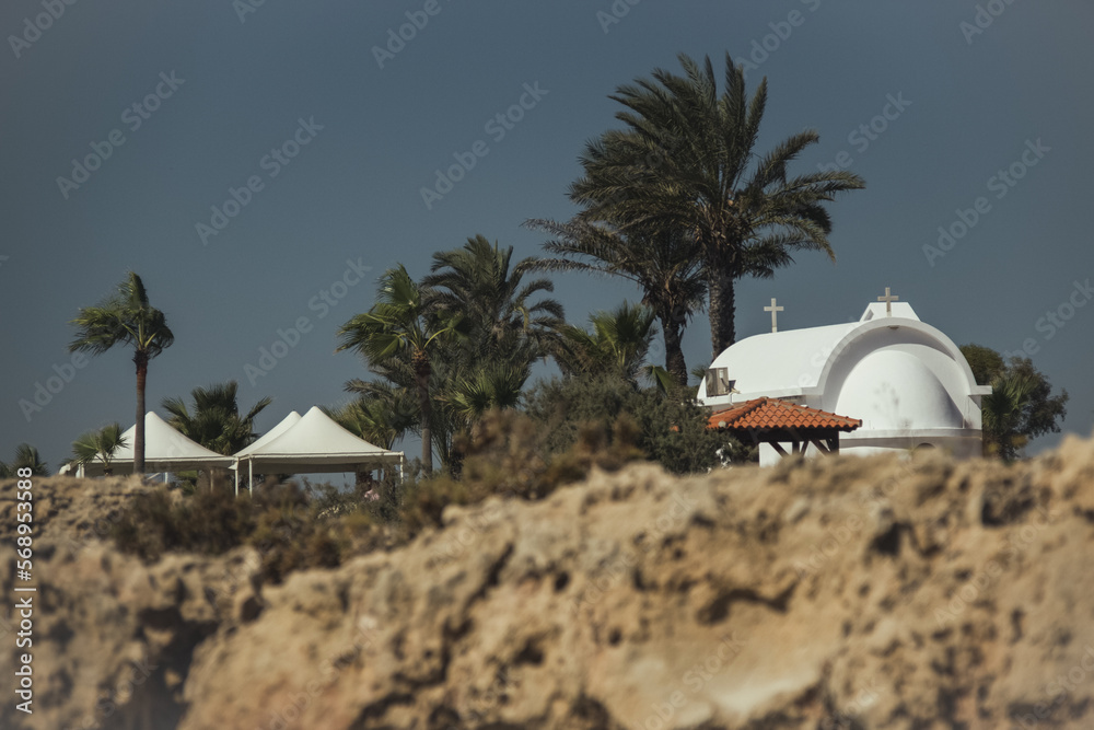 Naklejka premium Exterior view of small Catholic church painted in white in the city against palm trees and rocks. White church on a mediterranean island
