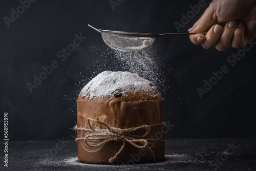 Traditional Italian Christmas cake Panettone tied with a jute thread on the black background. Pastry Chef sprinkles powdered sugar through a sieve on the fruit Easter cake with raisins.