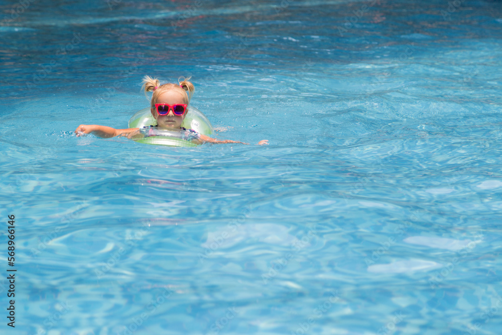 Happy little caucasian toddler girl with sunglasses smiling joyfully and enjoying summer vacations time in swimming pool in resort hotel. Kid with blonde hair wearing swimwear during summer holidays 