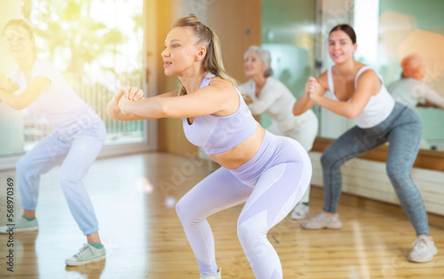 Smiling active women of different ages making squats during dancing training with coach in fitness studio
