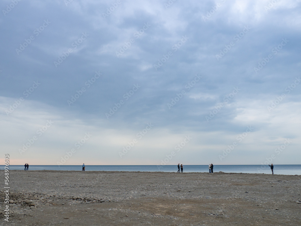 Small silhouettes of people on the shore. People in the distance stand and look at the sea. Overcast weather. Out of season at the